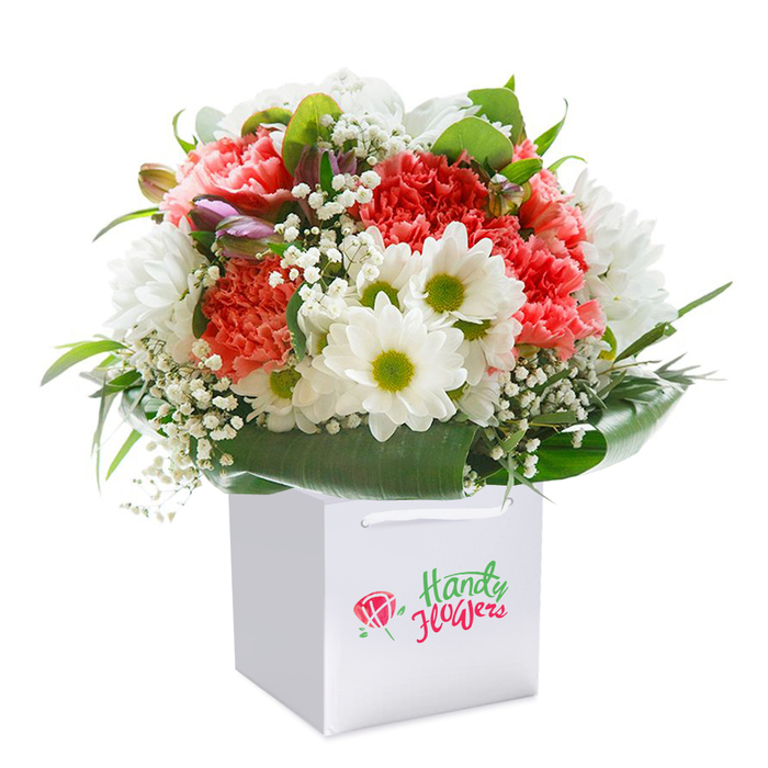 Bright and full of character, this expertly crafted bouquet rests elegantly in a white gift bag, ready to be delivered to a loved one across Kidbrooke. The composition brims with lush coral-pink and vivid red carnations-each petal delicately ruffled and vibrant, capturing movement and depth with their layered texture. Intertwined among the carnations, the crisp white petals and luminous green-yellow centres of daisies evoke the gentle charm of a walk through Kidbrooke's leafy spaces. Sprigs of airy white baby's breath (Gypsophila) drift throughout, their tiny blossoms providing a light, ethereal touch that softens the boldness of the main blooms. Tucked discreetly on one side, a few unopened buds display hints of purple and mauve, whispering of new beginnings and adding visual intrigue to the arrangement. The entire bouquet is embraced by deep green, naturally glossy foliage that cascades around the base, forming a luxuriant collar and highlighting the fullness of the design. The clean white bag, fitted with ribbons for comfortable carrying, grounds the composition in practicality and understated elegance-perfect for hand-delivery anywhere from Shooters Hill to Blackheath. Photographed in brilliant studio light with no distractions, every detail from the gentle shimmer on the leaves to the interplay of colours in the blooms is showcased. Evoking feelings of care, freshness, and joy, this bouquet is a testament to Kidbrooke's blend of classic beauty and modern sensibility, brought to life by local artisans.