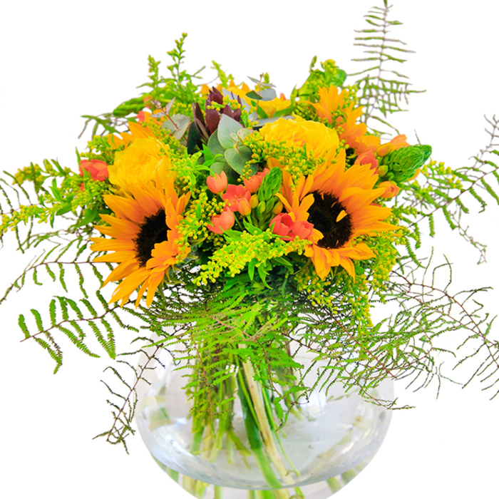 Radiant and full of life, this hand-crafted floral arrangement captures the essence of seasonal beauty so familiar to Kidbrooke. Dominated by resplendent sunflowers with bold yellow petals and striking brown centers, the bouquet is intertwined with golden spirals of garden roses. Each rose's closely-layered petals adds a classical, textural richness, their buttery scent floating lightly in the air. Tucked amongst the sunflowers and roses, clusters of luminous red Hypericum berries evoke playful bursts of colour, reminiscent of berry-laden hedgerows bordering the local paths. Feathery fronds and a profusion of greenery-some delicate, others more structured-spill over the edges, echoing the abundance of Plantagenet Park's summertime foliage. Light green sprigs, possibly solidago, scatter tiny yellow blossoms throughout, enhancing the feeling of a landscape in full bloom. Silvery eucalyptus leaves weave through the bouquet, lending a subtle musk and a frosted palette touch, perfect for both modern and rustic tastes. The vase itself, a pristine clear sphere, reveals the arrangement's architecture-the intertwining stems and shimmering water evoking freshness. Bathed in gentle daylight, the entire composition feels alive and celebratory, ideal for birthdays, local milestones, or simply injecting a sense of joy into a Kidbrooke home. This piece, reflecting the creativity of Florist Kidbrooke, distils both the natural beauty and spirited warmth of the SE3 community in every carefully chosen bloom.