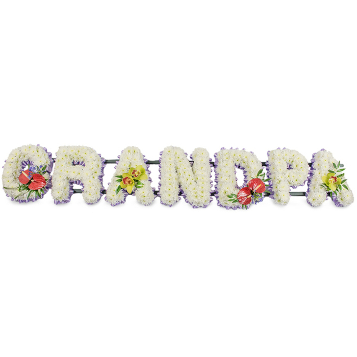 A striking, personalised funeral flower display spells out "GRANDPA" in bold capital letters, designed as a long, horizontal arrangement set against a clean white backdrop so every petal can be imagined clearly. Each letter is shaped from densely massed white chrysanthemums, the small, daisy-like blooms arranged so tightly that no gaps show, creating a smooth, cushiony surface that feels calm and reassuring. The white flowers appear almost luminous against a slender band of pale lilac or lavender ribbon that borders the base of each letter, adding gentle definition and a touch of quiet elegance. Subtle green support bars run beneath the word, giving the structure strength while keeping the focus firmly on the flowers themselves. Carefully placed sprays introduce colour and personality: on the far-left "G" and the central "D," clusters of rich red anthuriums are arranged with their characteristic heart-shaped, glossy spathes and upright yellow spadices. These deep crimson tones, framed by dark green leaves and tiny hints of purple filler flowers, suggest deep affection, courage, and heartfelt remembrance. On the first "A" and the final "A," graceful yellow-green orchids with slender petals and patterned throats rest among lush foliage, bringing a fresh, modern lightness that lifts the design. The tribute feels both traditional and contemporary, the type of bespoke "Grandpa" lettering often chosen by families in Kidbrooke for funerals at local chapels or crematoriums, or for placing at a graveside after a quiet walk from Kidbrooke Village. It speaks of a grandfather cherished for his warmth and constancy, and of a family wanting a floral memorial that captures both their grief and their gratitude in a single, beautifully crafted word.